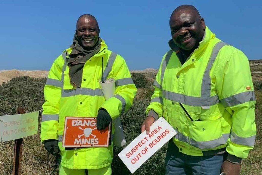 ELLITORAL_338128 |  Fotografía tomada de FITV, primera estación de televisión local de las Islas Malvinas. Shame Mapulanga y Thomas Hlaisi, desmineros procedentes de Zimbabwe, muestran algunos de los carteles que advertían sobre la presencia de minas bajo tierra.