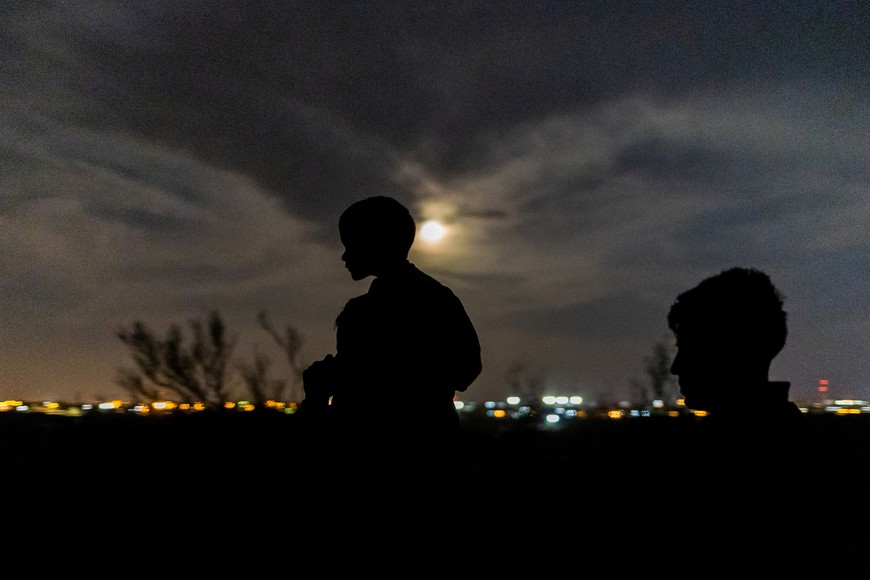 The Sturgeon moon rises past city lights of Ciudad Miguel Aleman, Mexico in the background as Felix, an asylum seeking migrant from Peru, carries his three year old son Young along a dirt trail after crossing the Rio Grande river into the United States from Mexico in Roma, Texas, U.S., August 11, 2022. REUTERS/Adrees Latif