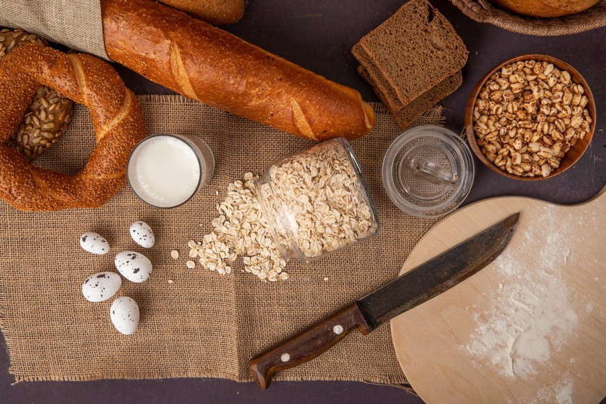 top view of breads as bagel rye and baguette with milk oat-flakes eggs corns and knife on sackcloth on maroon background