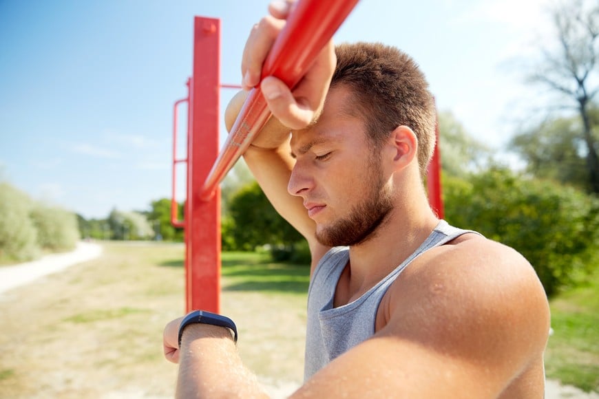fitness, sport, training and lifestyle concept - young man looking at heart-rate watch bracelet and exercising on horizontal bar outdoors